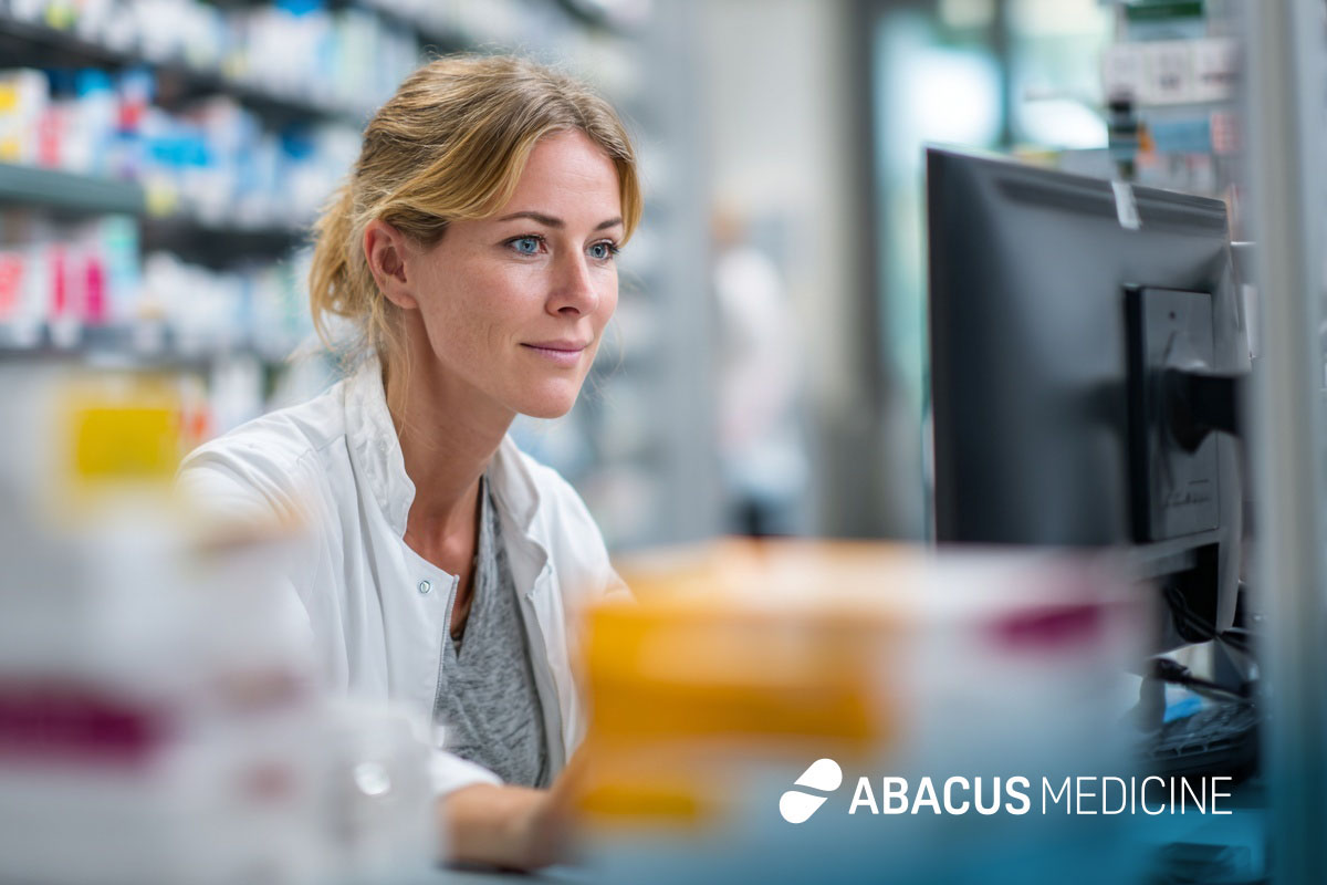 Eine blonde Apothekerin im weißen Kittel blickt lächelnd auf einen Computerbildschirm vor vollen Medikamentenregalen. Unten rechts befindet sich das Logo von Abacus Medicine.