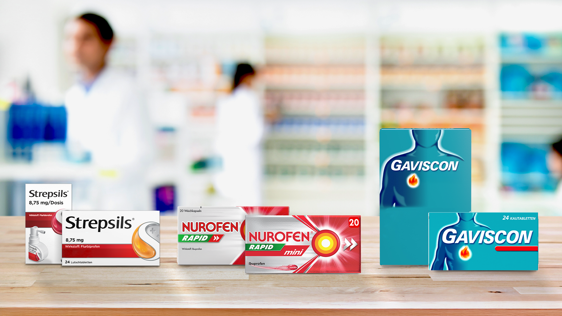 Medikamentenpackungen der Marken Strepsils, Nurofen und Gaviscon auf einem Holztresen vor dem unscharfen Hintergrund einer Apotheke.