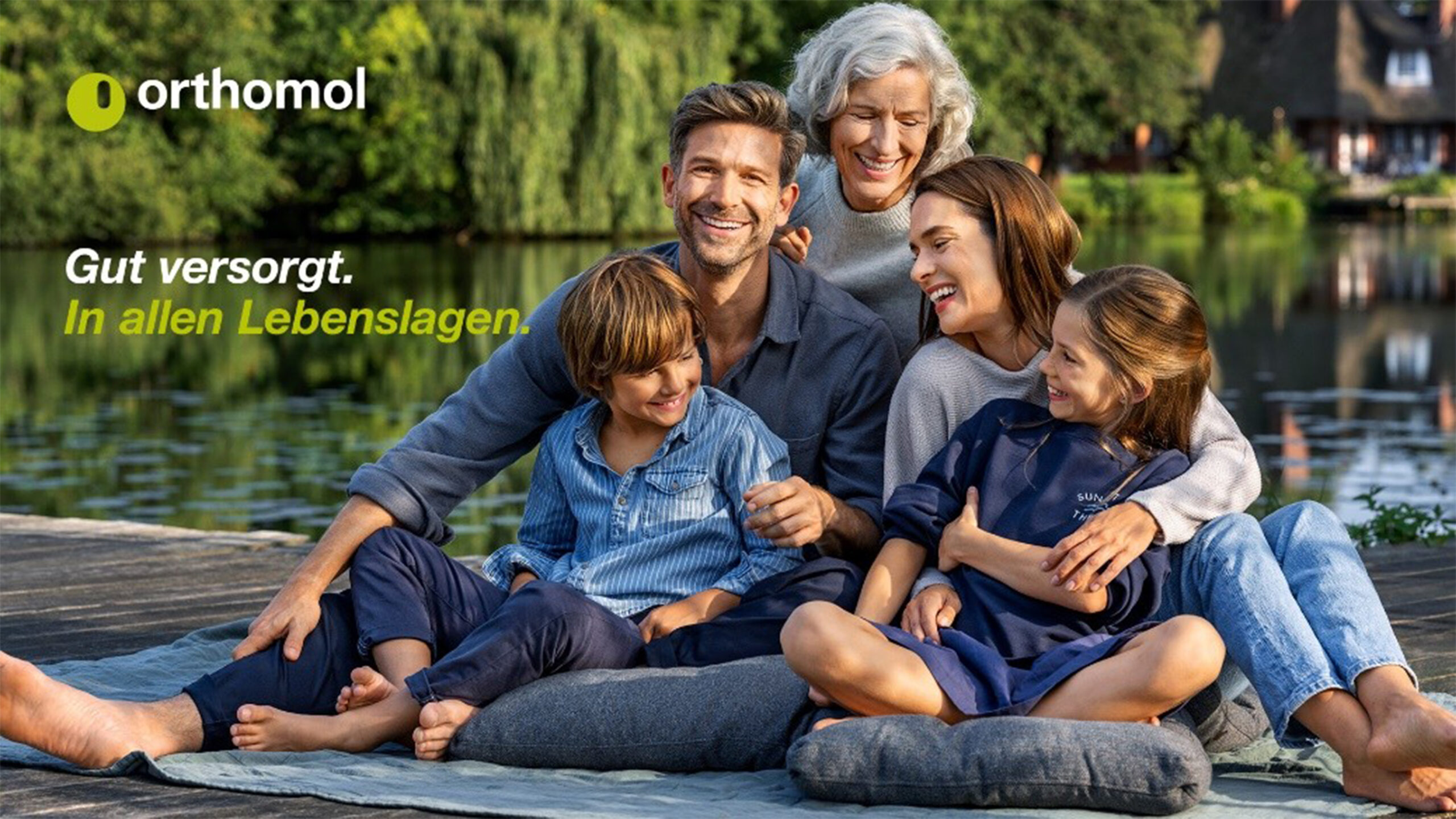Fünfköpfige Familie, bestehend aus Großmutter, Vater, Mutter und zwei Kindern liegt sich, auf einem Steg am Fluss sitzend, in den Armen und lacht.