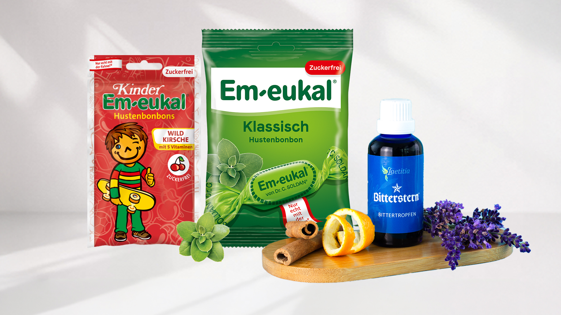 Produktarrangement vor hellem Hintergrund: Eine rote Packung 'Kinder Em-eukal Wildkirsche' mit Jungen-Motiv, eine grüne Packung 'Em-eukal Klassisch' und eine blaue Flasche 'Bitterstern Bittertropfen' auf einem Holzbrettchen mit Kräutern, Zimtstangen und Orangenschalen.