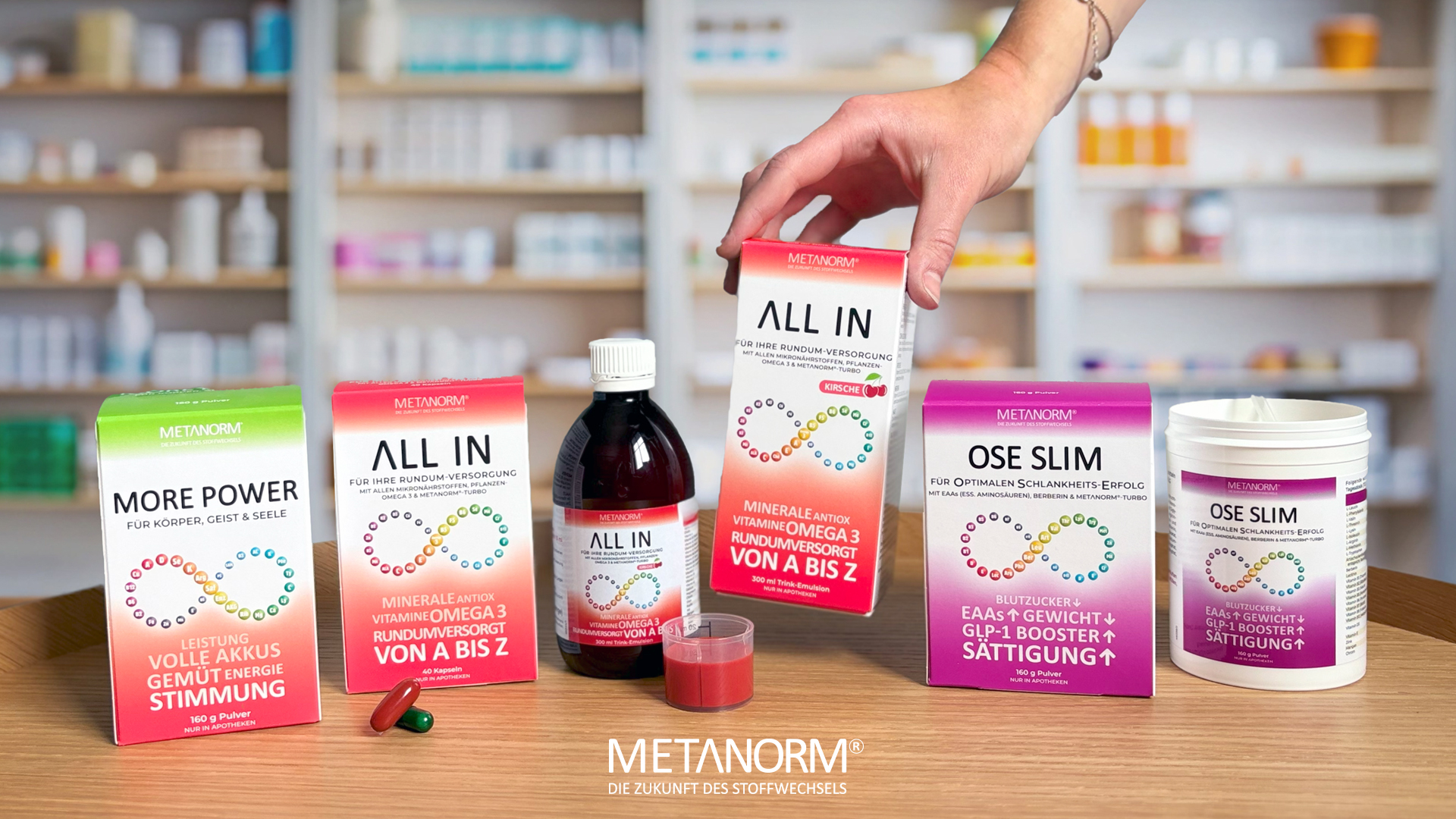 Produkte der METANORM Serie präsentiert auf einem Tisch: ,,MORE POWER Pulver", ,,ALL IN Kapseln und Trink-Emulsion" und „OSE SLIM Pulver". Eine Hand greift nach der „ALL IN Trink-Emulsion".