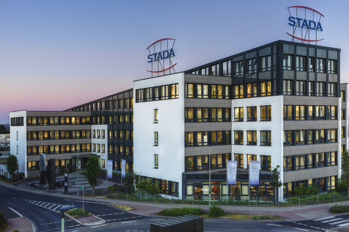 Außenansicht der STADA-Zentrale bei Abenddämmerung; modernes Bürogebäude mit großen Fenstern, beleuchteten Innenräumen und zwei STADA-Logos auf dem Dach; davor Fahnenmasten mit Firmenfahnen und eine gepflegte Straßenkreuzung