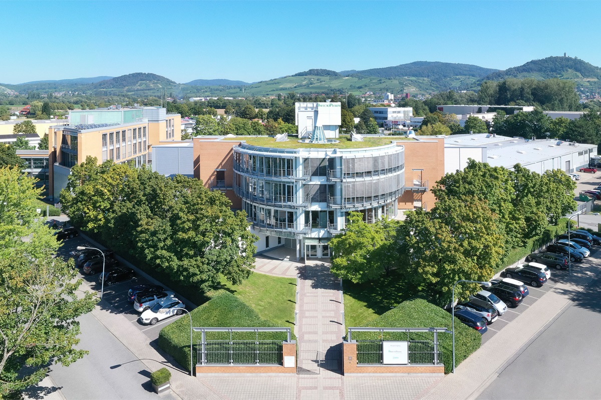 Luftaufnahme des modernen InfectoPharm-Unternehmensgebäudes: Ein halbrunder, mehrstöckiger Glasbau mit begrüntem Dach, umgeben von Bäumen, Parkplätzen und einer gepflegten Außenanlage vor einer Hügellandschaft im Hintergrund.